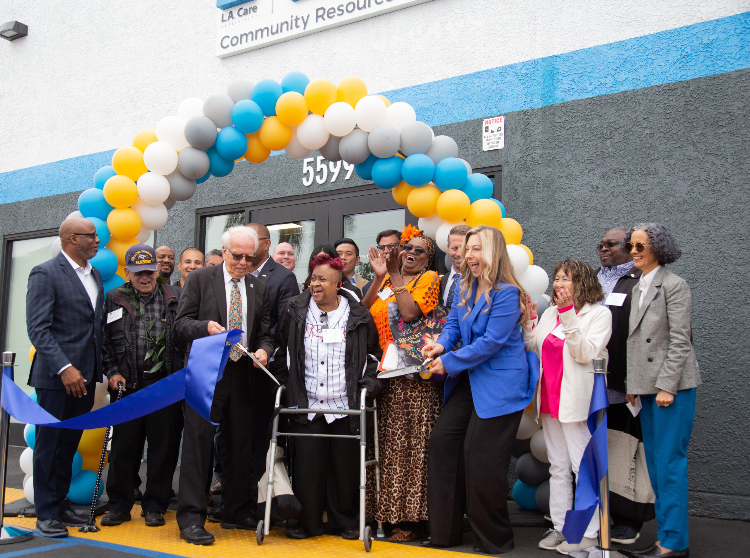 A group of people surrounded by an arch of balloons, smile while cutting a ribbon.