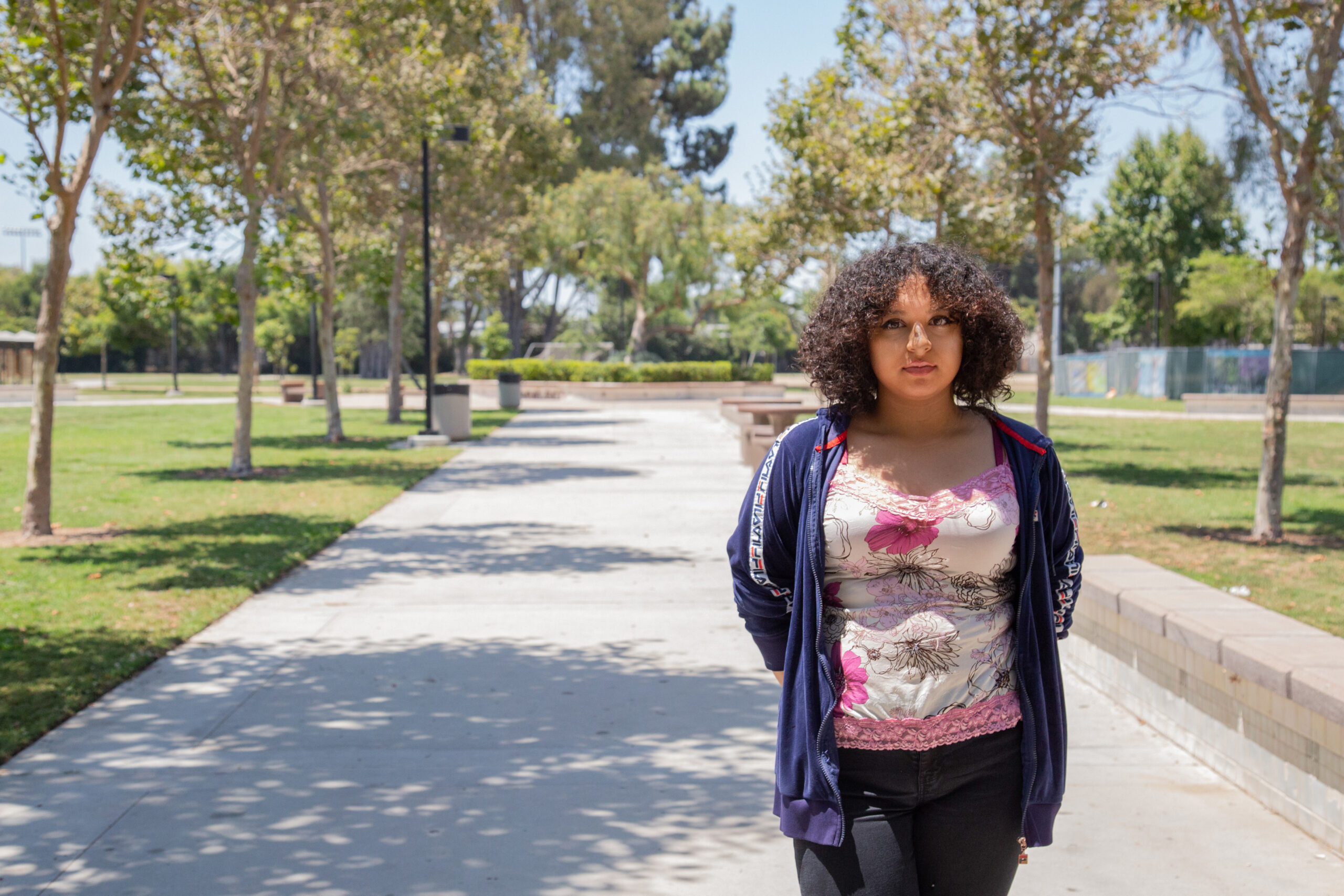 A girl wearing a flower-printed shirt and a blue jacket stands to the right of a sidewalk which is visible behind her and is lined with trees on either side.