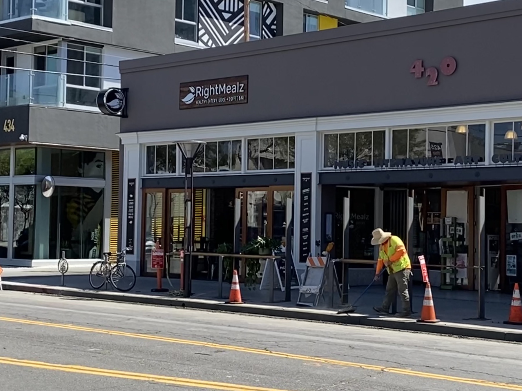 Traffic cones and a construction worker are seen on the sidewalk of a building that says "RightMealz" and "420."