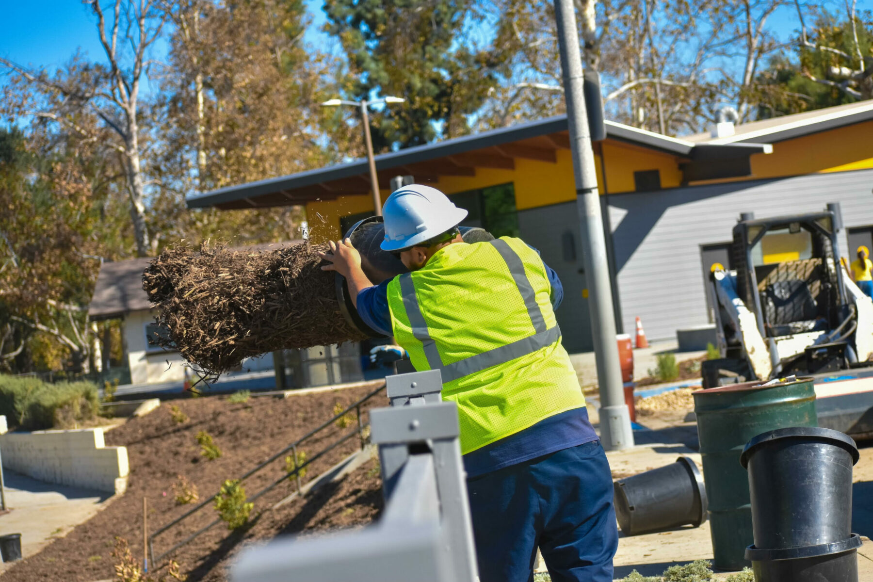 A man in a neon yellow vest and white construction hat tosses soil into a garden.