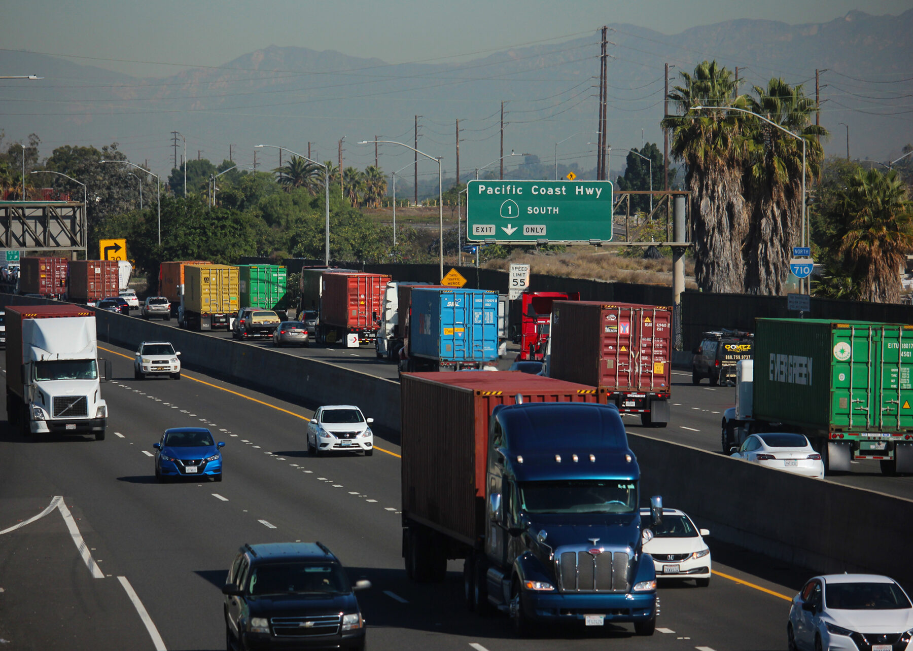 A line of semi-trucks travel along a highway with mountains in the background.