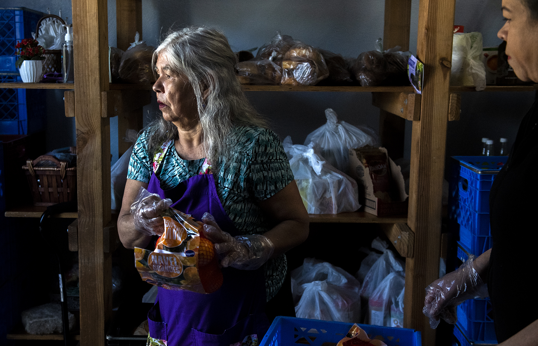 A woman wearing a blue apron and clear plastic gloves holds a bag of oranges.