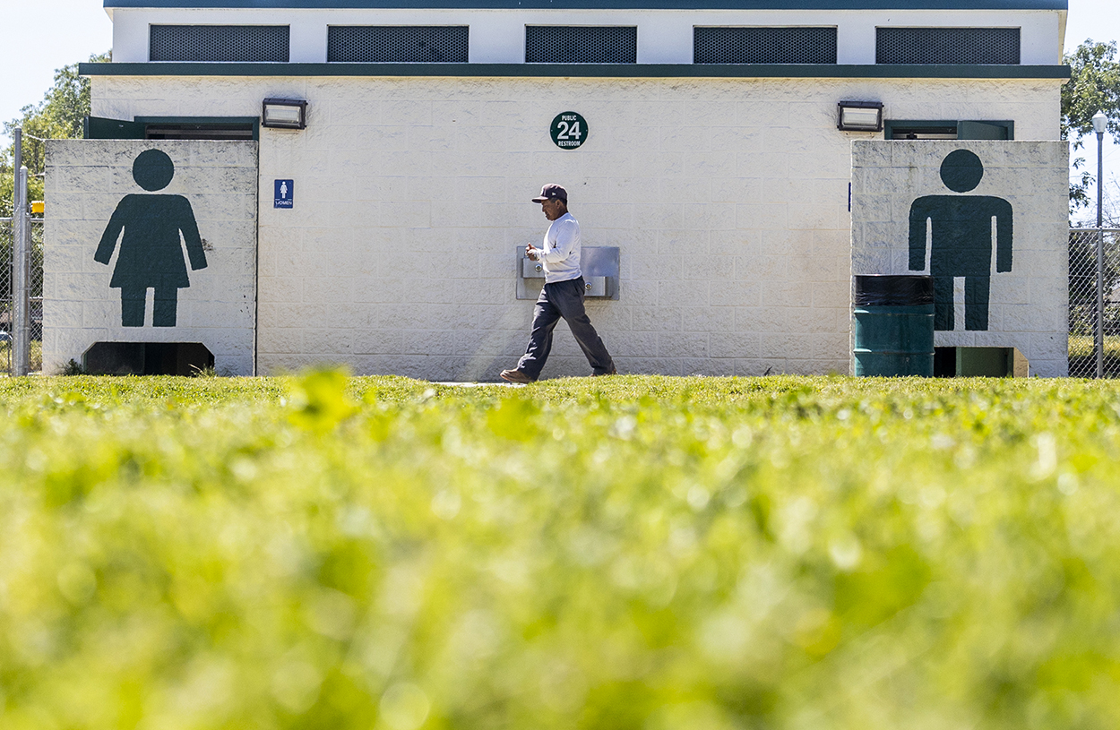 A man in a white shirt, jeans and baseball cap walks by an outdoor restroom building.