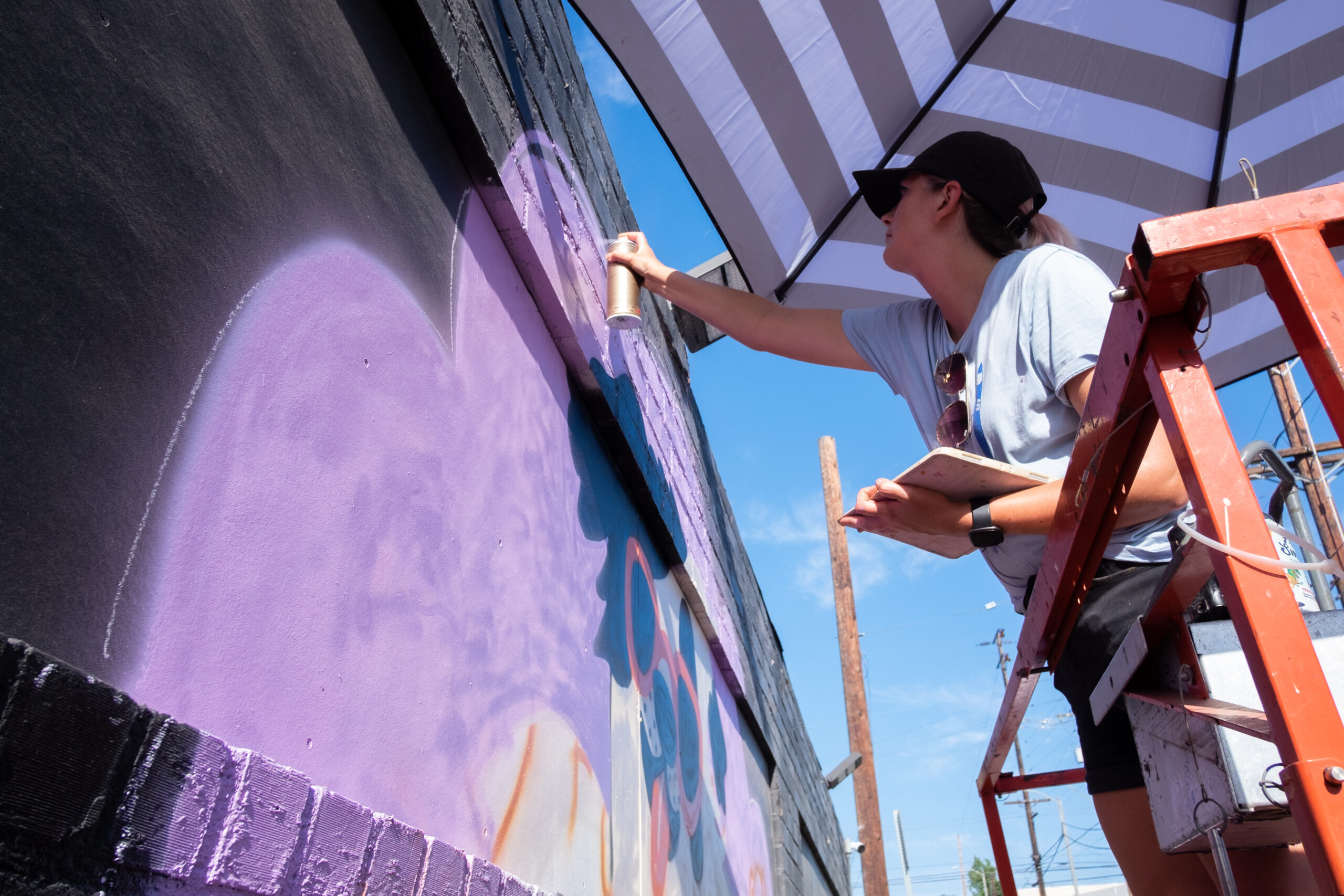 An artist stands on a ladder and sprays a purple wall. A striped umbrella is in the background along with a blue sky.