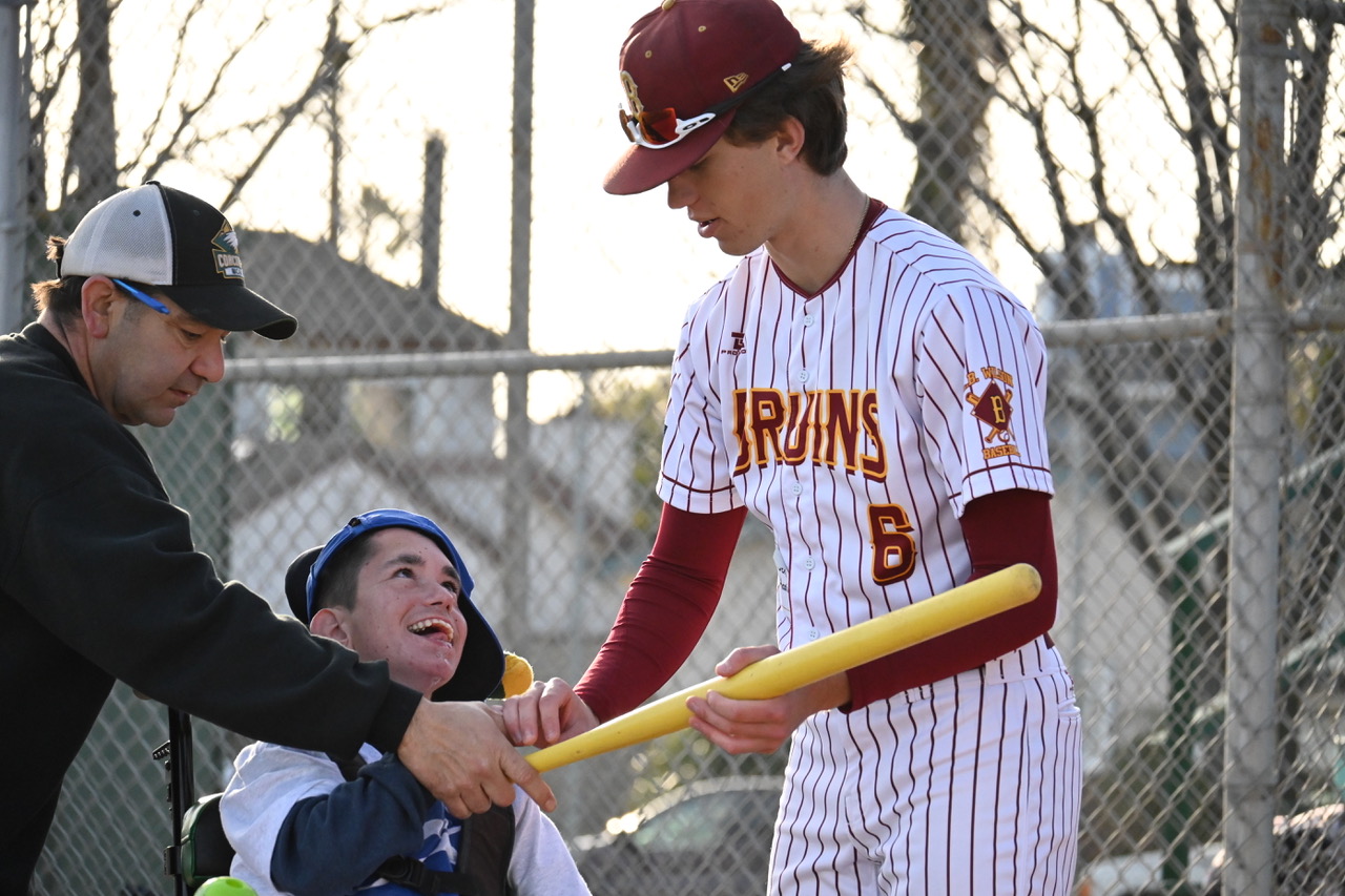 A person in a baseball uniform helps a person in a wheelchair hold a baseball bat.