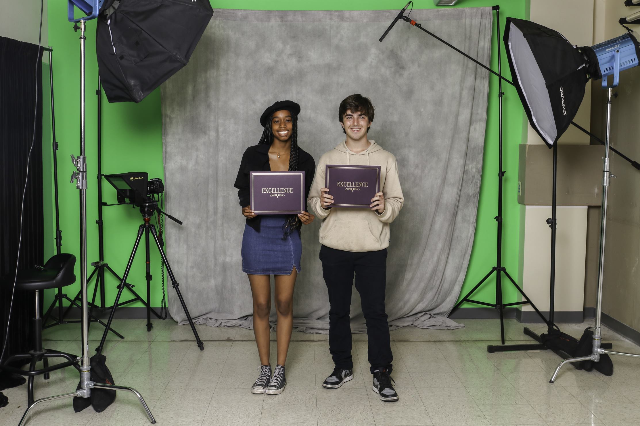 Two students stand in a photography studio holding up certificates that say "Excellence."