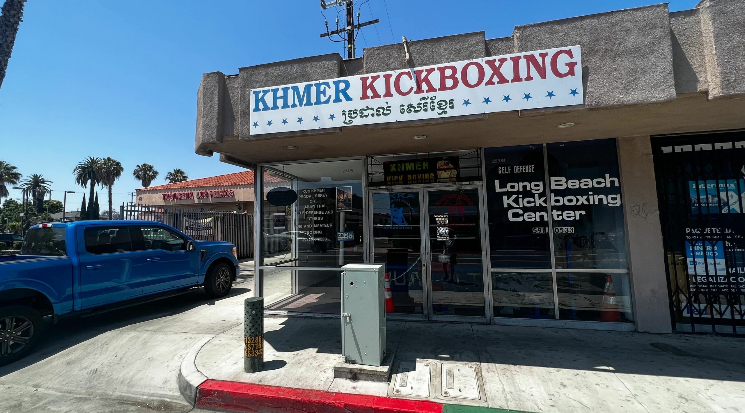 A building with the sign "Khmer Kickboxing" at the top, a blue car is parked next to it.