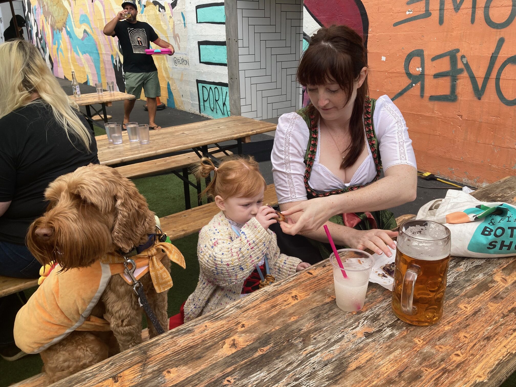 A woman at a picnic table gives a piece of food to her small daughter, who is sitting next to a large dog/