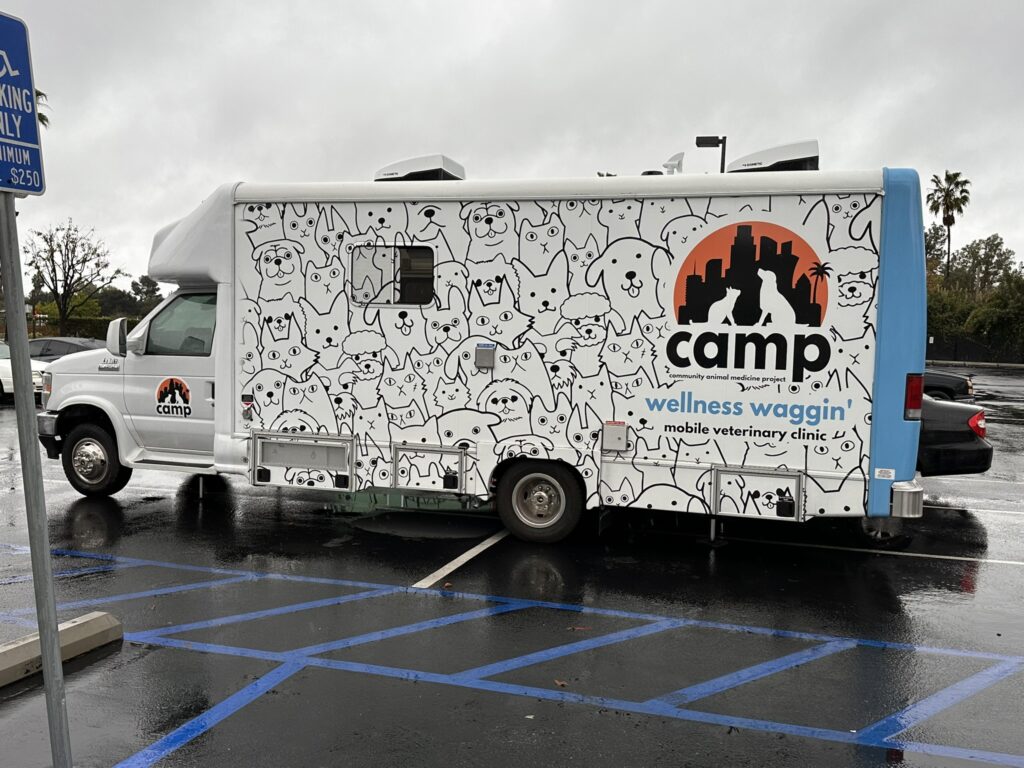 Large van with the word "camp" on it and "wellness waggin'" with line drawings of cats and dogs sits in a rainy parking lot.