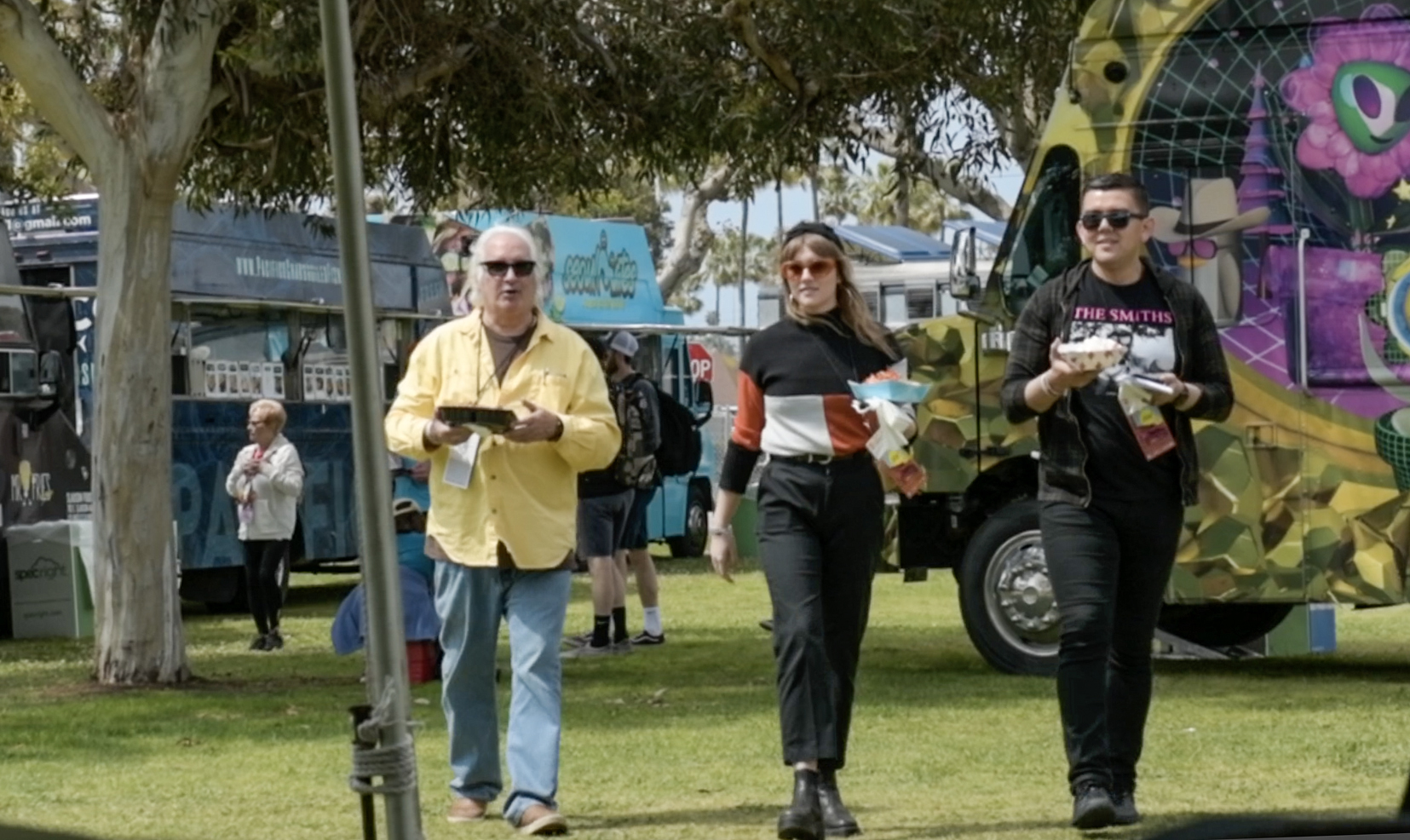 Three people walking while holding food with food trucks in the background.
