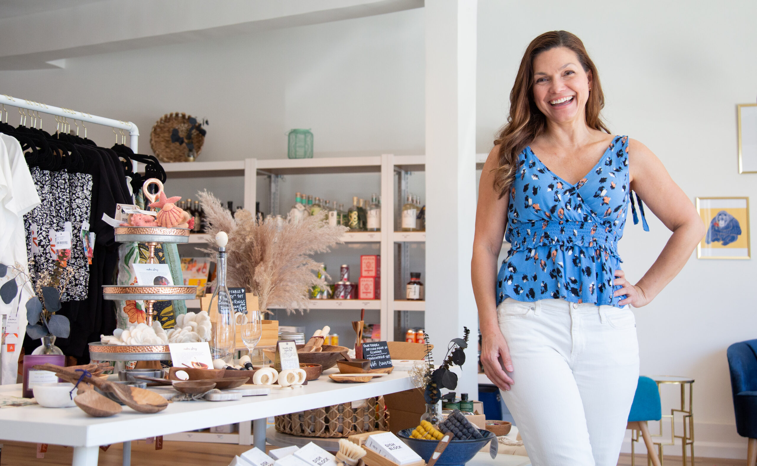 A woman in a blue shirt and white pants smiles with one hand on her hip. A table with items, a shelf behind her with bottles, and a clothing rack are visible.
