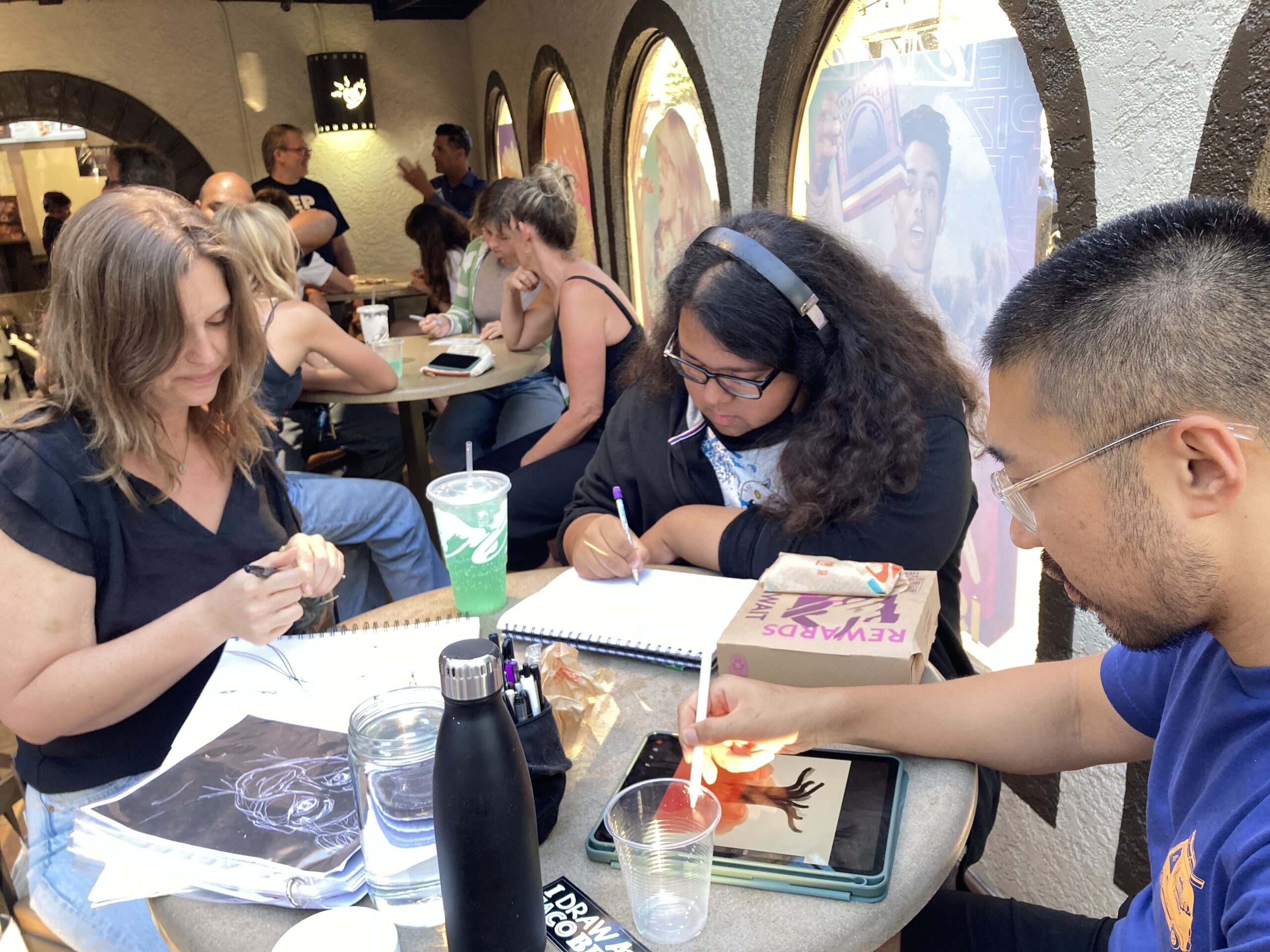 Three people sit at a table in a Taco Bell restaurant drawing pictures.