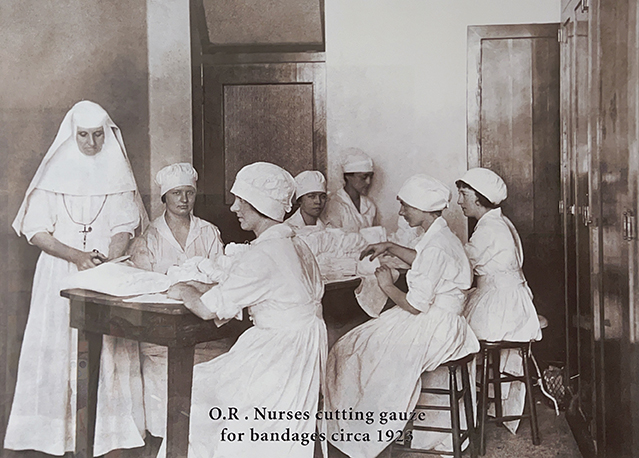 A group of women in white sit at a table working with cloth in a black-and-white photo that says, "O.R. nurses cutting gauze for bandages circa 1923."