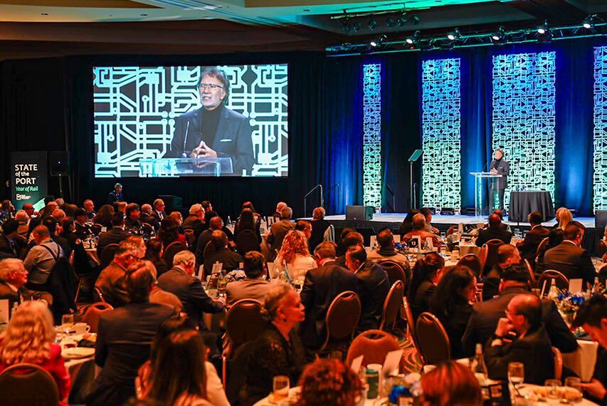 A man in business attire on a stage speaks to a crowd seated at tables.